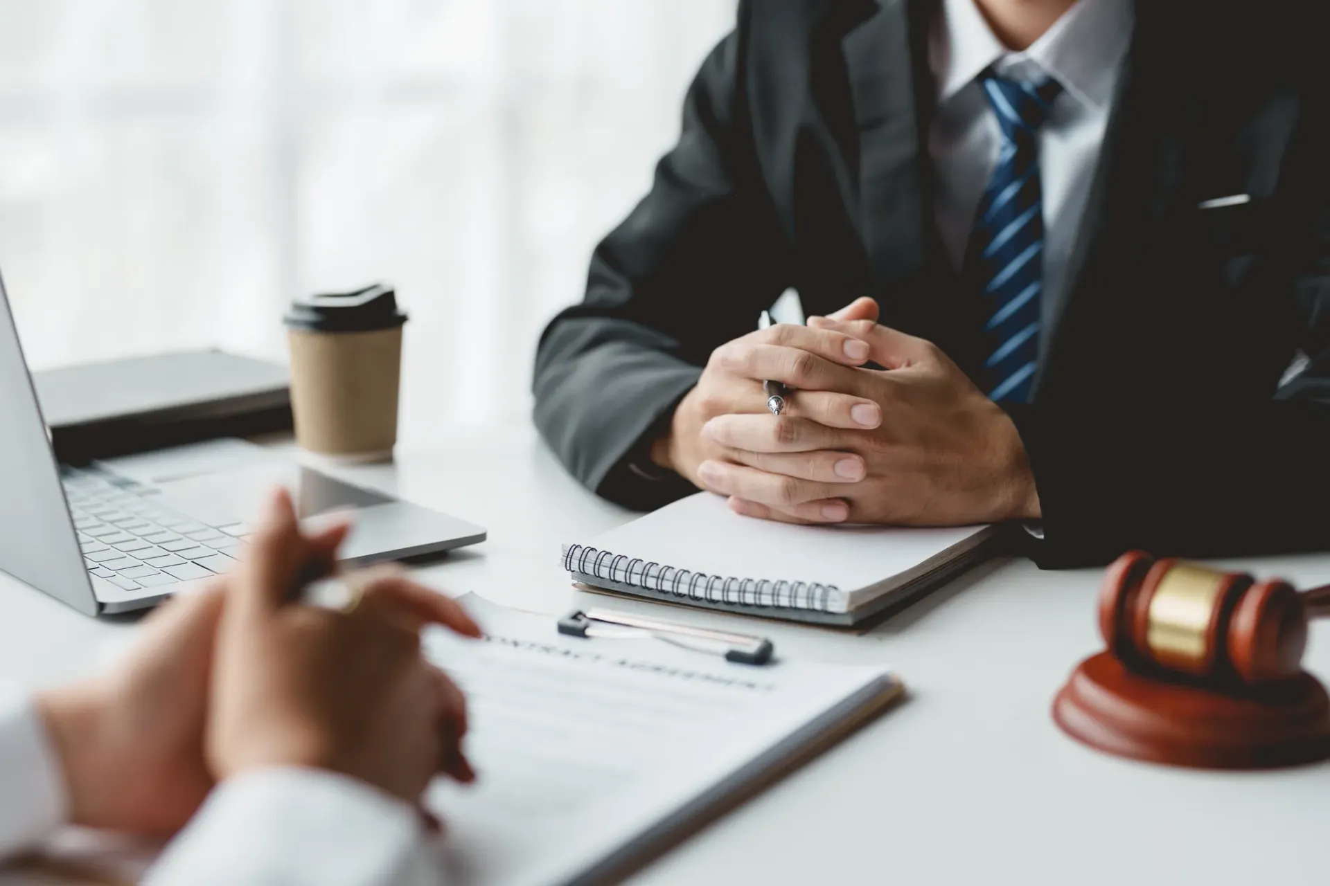 Business meeting with two people across a desk, laptop, coffee cup, notebook, and gavel in view