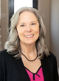 Smiling woman with gray hair wearing a black cardigan and pink top against a neutral background
