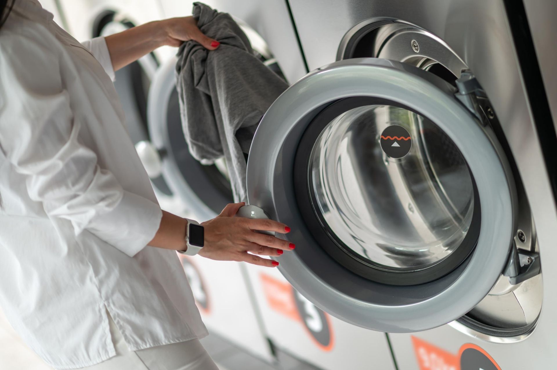 Person adjusting a front-loading washing machine, with clothes in hand and laundry appliances visible.