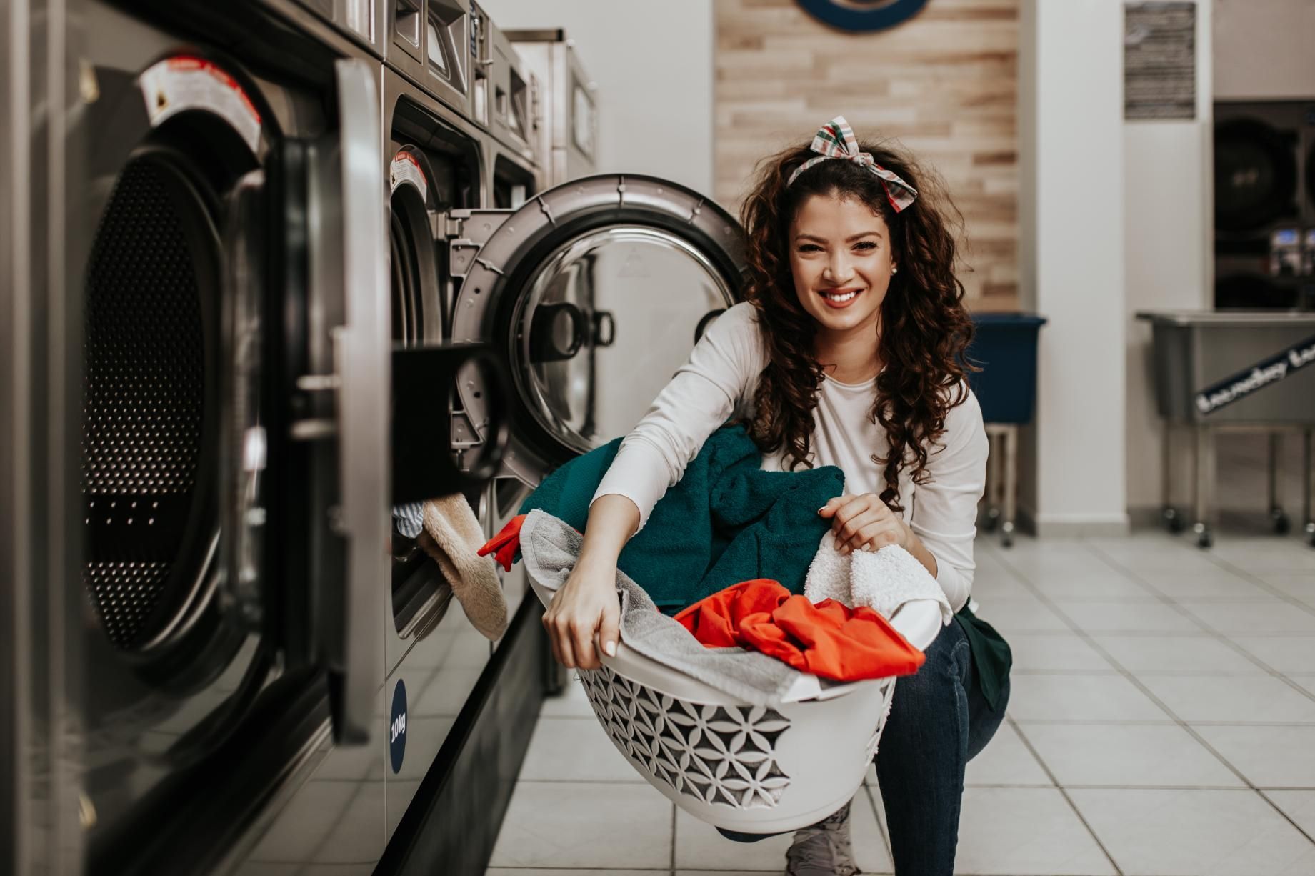 Woman crouches with a laundry basket of red clothes in a laundromat, smiling beside washing machines.