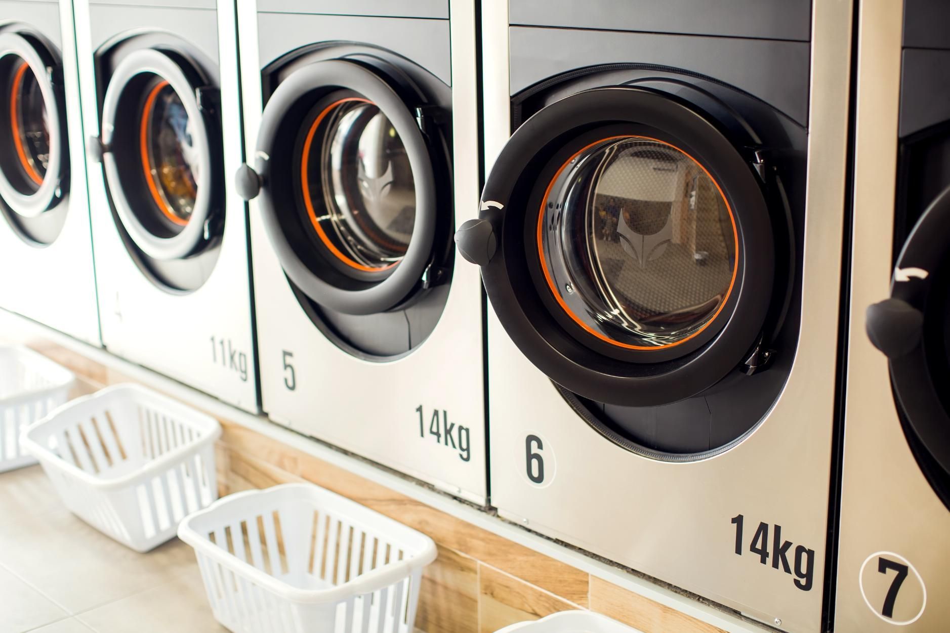 Row of front-loading washing machines in a laundromat with laundry baskets on the floor