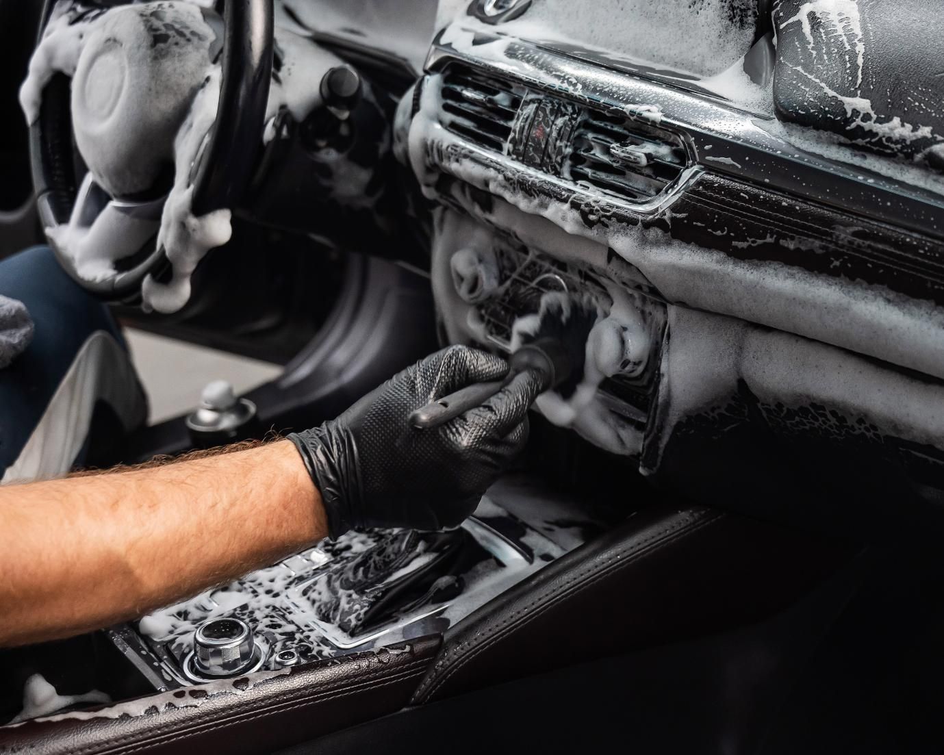 Gloved hand cleaning a car dashboard and center console with foamy soap.