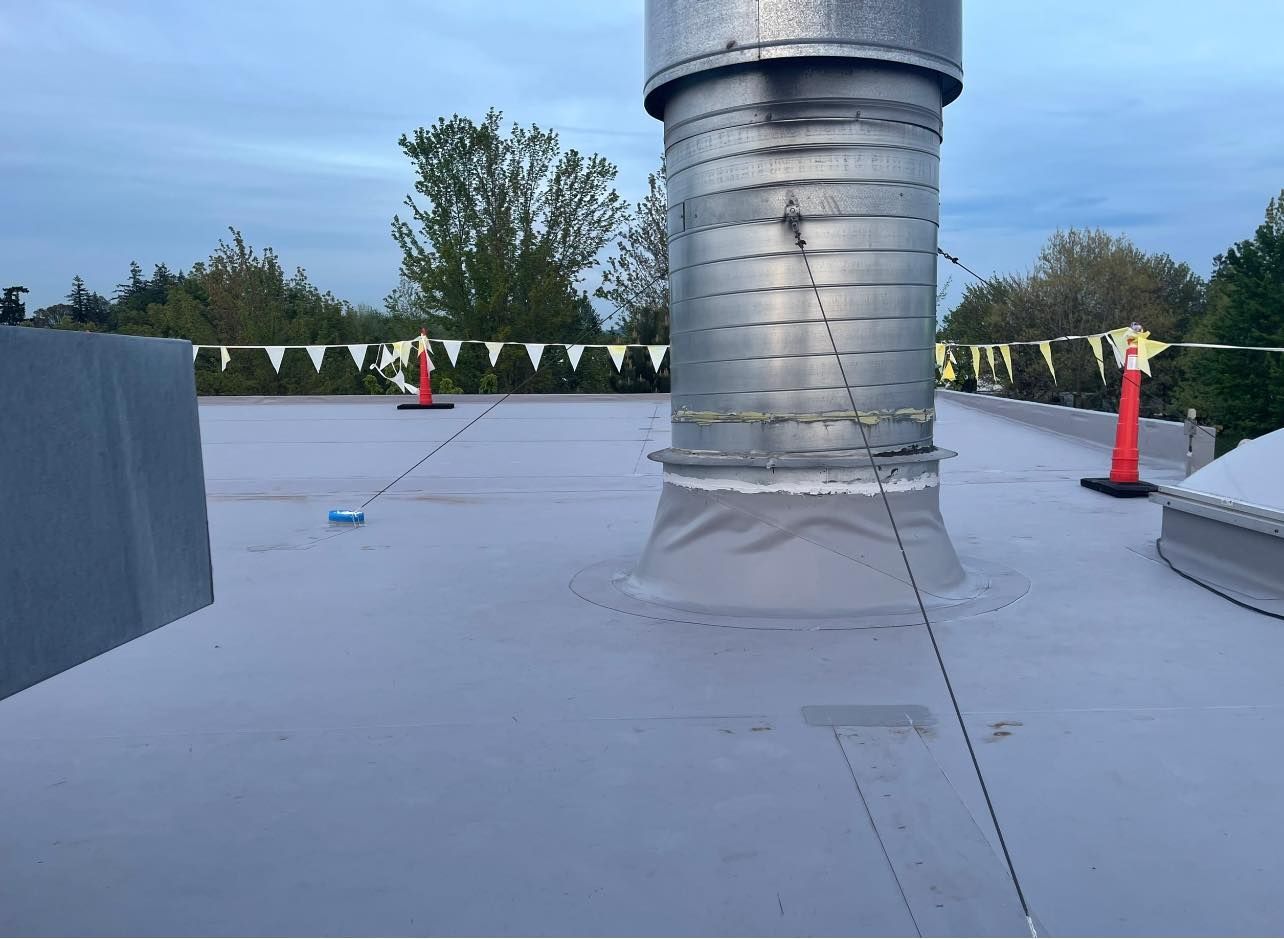Flat rooftop with gray roofing, a large silver vent, and trees under a cloudy sky