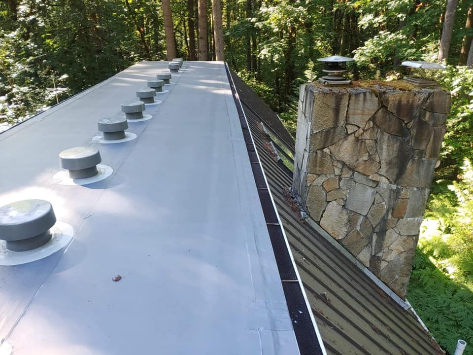 Rooftop with white metal panels and vents beside a stone chimney in a wooded area