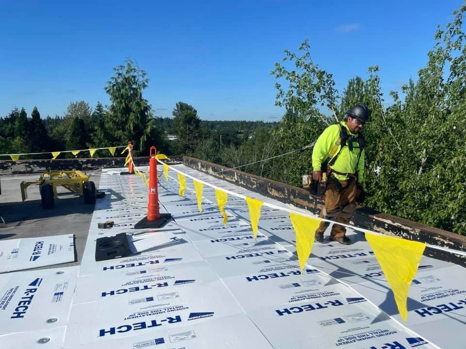 Worker in neon safety gear on a rooftop, installing yellow caution tape beside orange cones under a blue sky