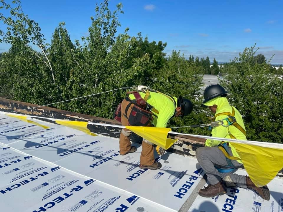 Two workers in safety gear installing roofing underlayment on a sloped roof near trees and blue sky