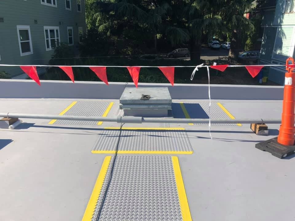 Rooftop walkway with yellow safety markings, red caution flags, and an orange traffic cone near a hatch.