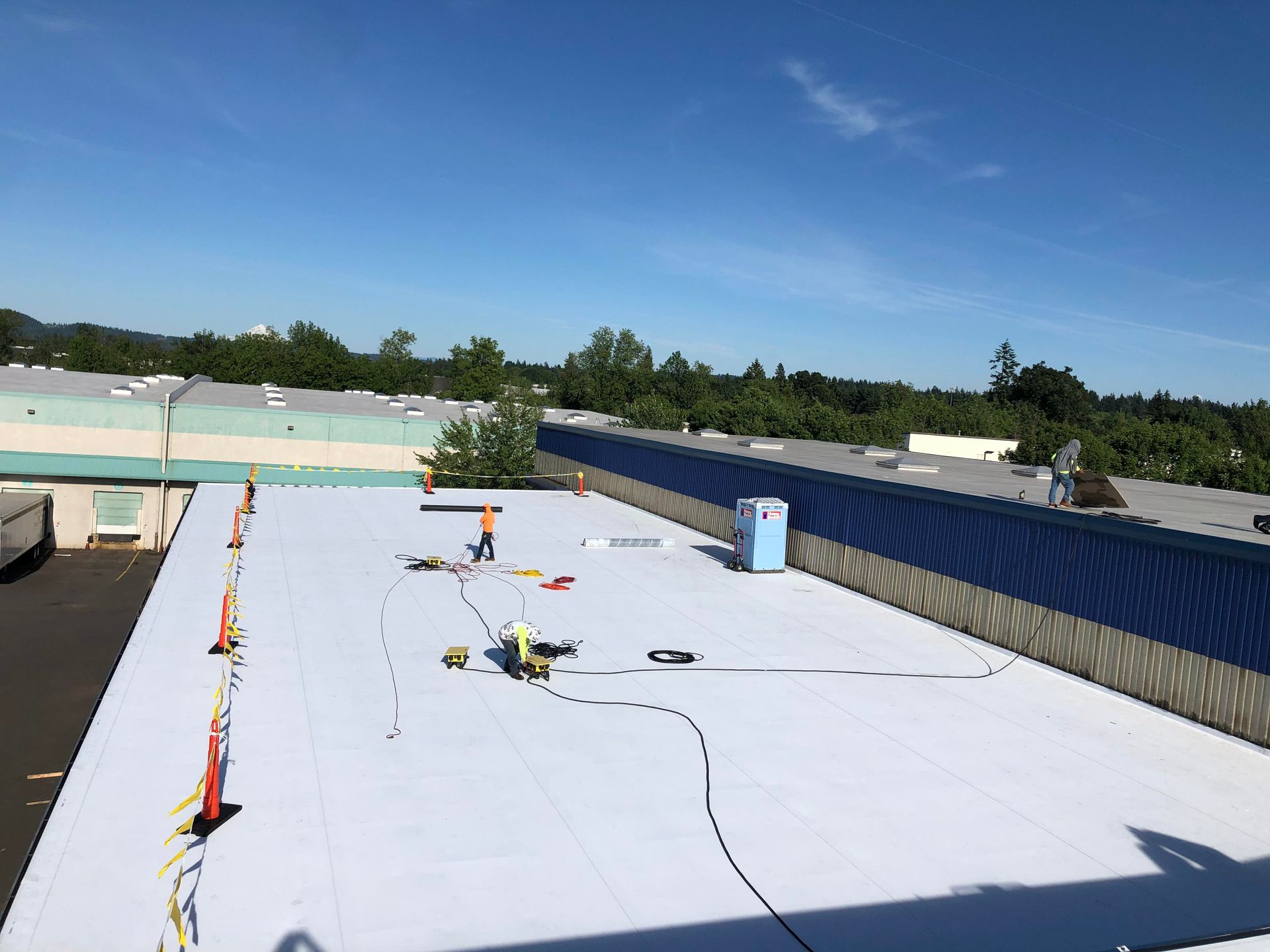 Flat white rooftop under a blue sky, with scattered equipment, orange cones, and a blue vent line.
