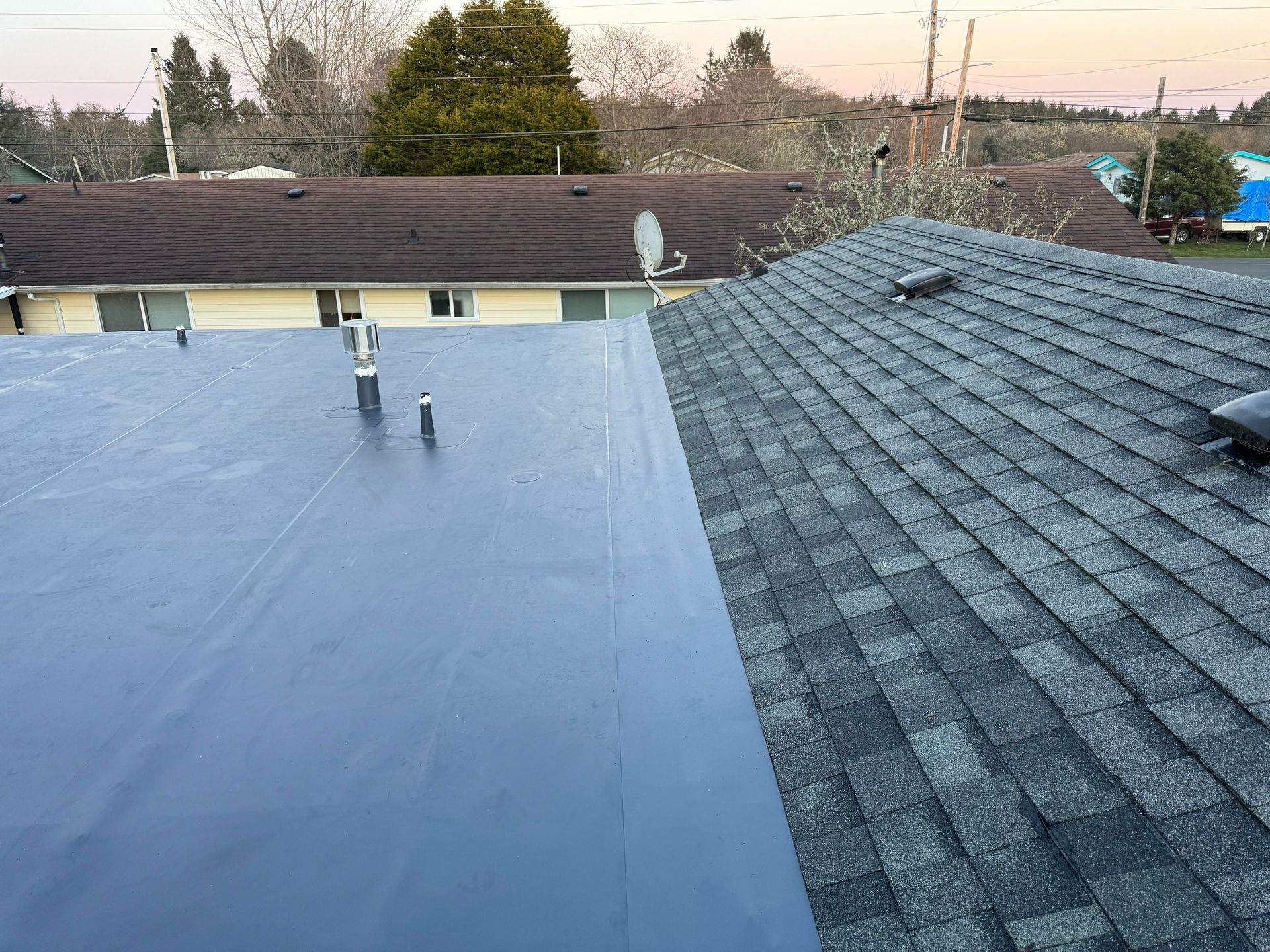 Flat rooftop with blue membrane next to gray shingle roof at sunset; trees and utility poles in background