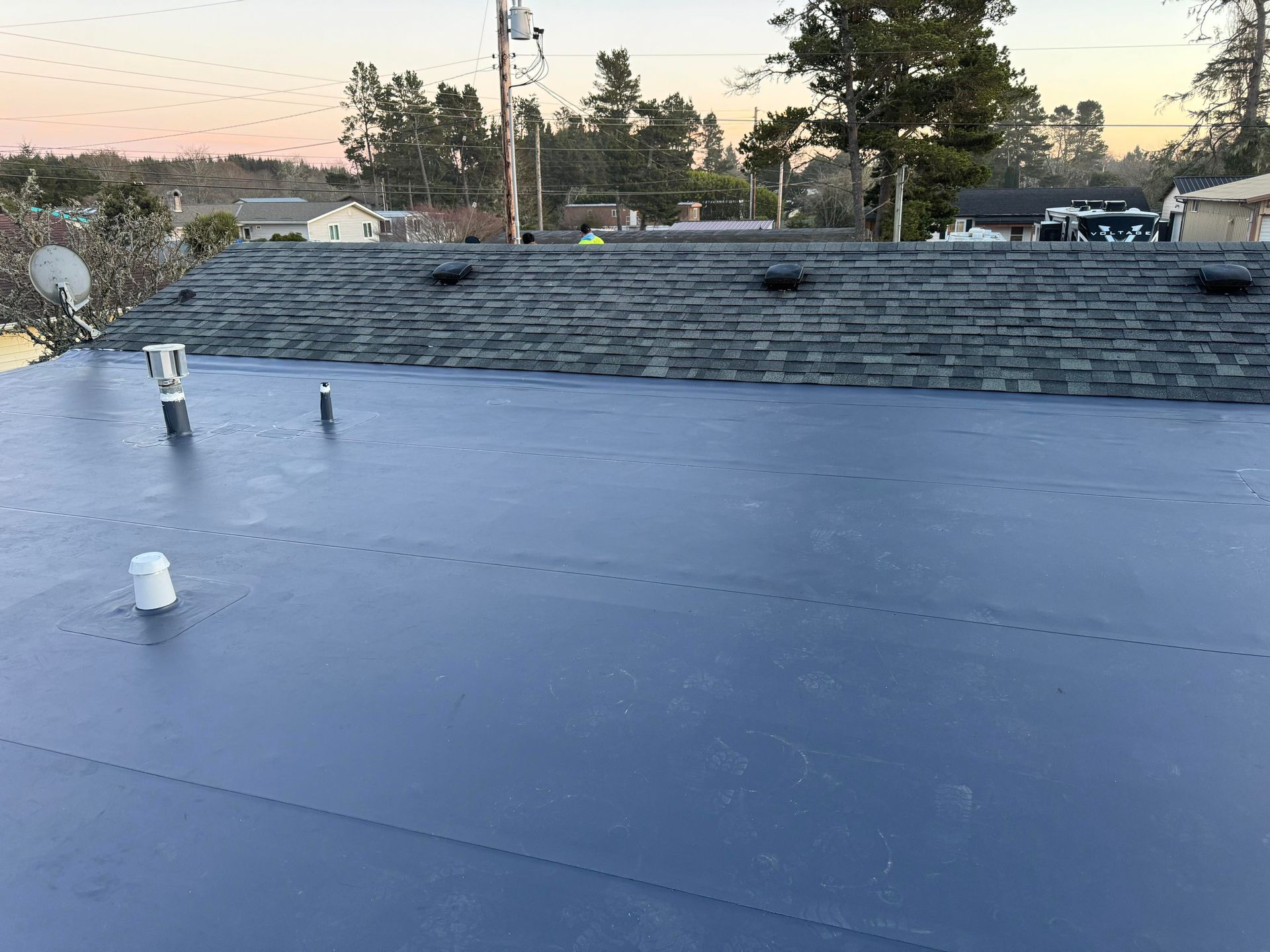 Flat blue roof with vents and a low stone wall at sunset, with trees and houses in the background