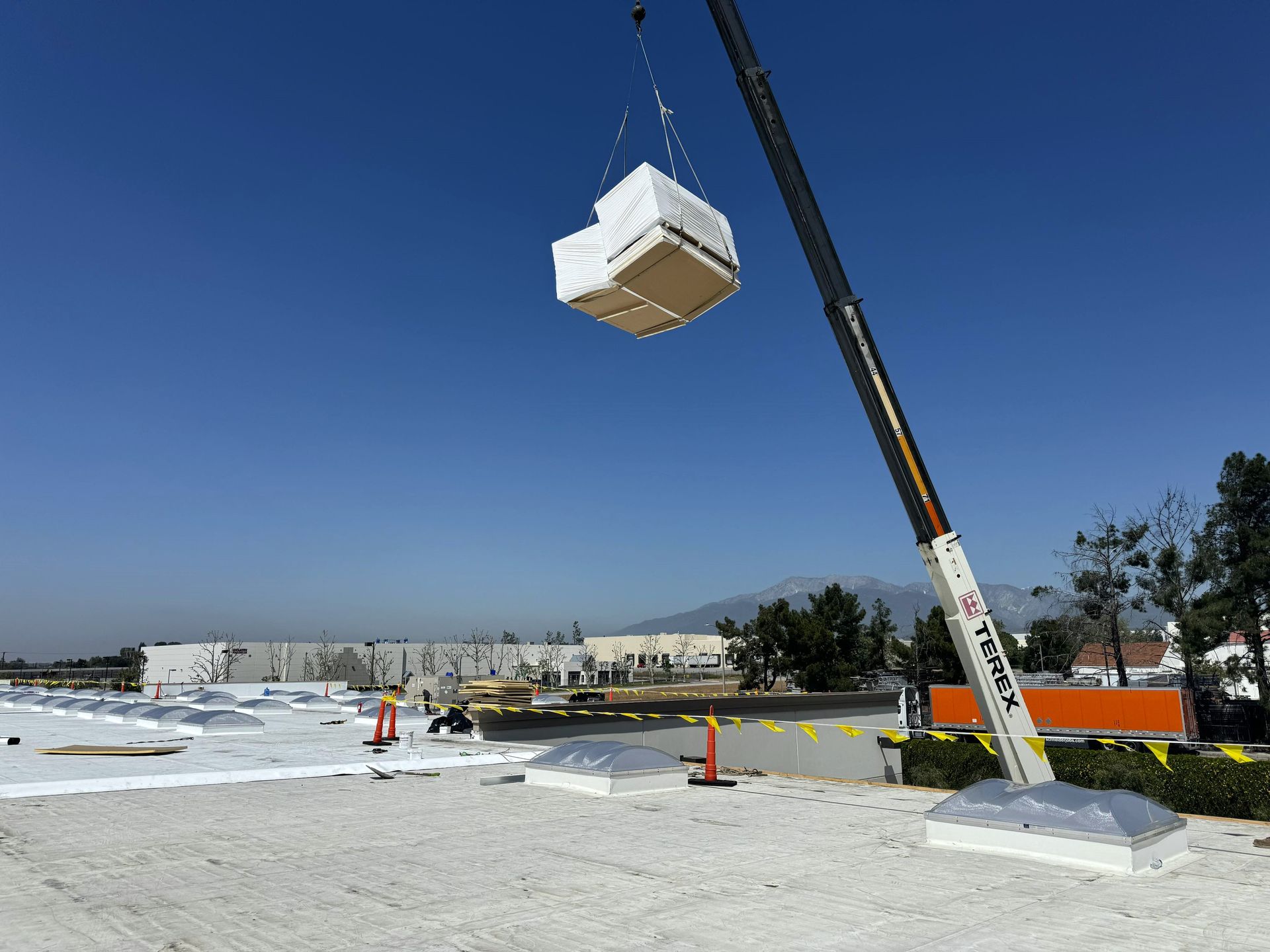 Crane lifting a large white block over a commercial roofing construction site under a clear blue sky