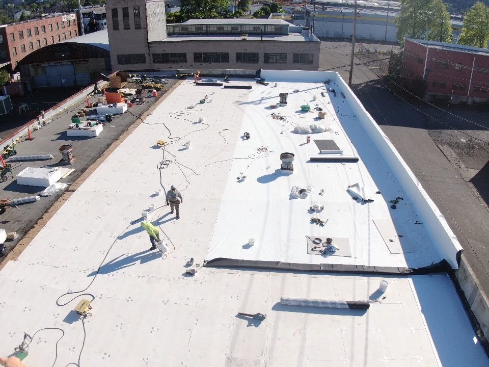 Rooftop with a white membrane being installed, workers and equipment visible on a sunny day.