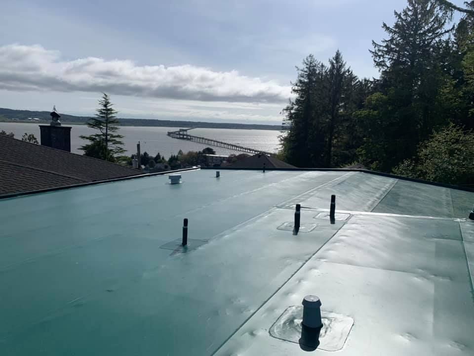 Green rooftop with vents overlooking trees, houses, and a distant body of water under cloudy skies