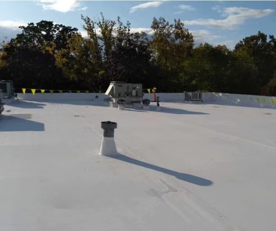 Flat white rooftop with vents and equipment under a blue sky, backed by trees.