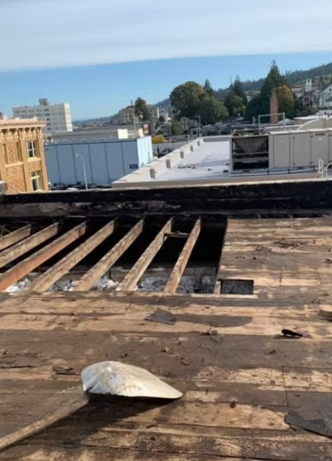 Rooftop with exposed wooden beams and weathered roofing, overlooking buildings and a distant blue sky.