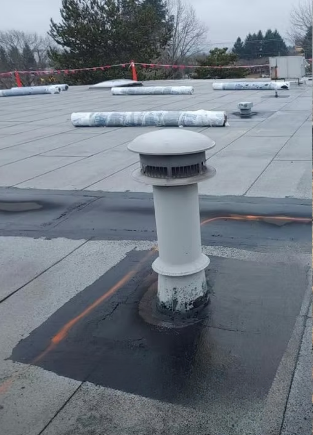 Rooftop vent pipe with white cap on a flat roof, surrounded by HVAC units and overcast sky