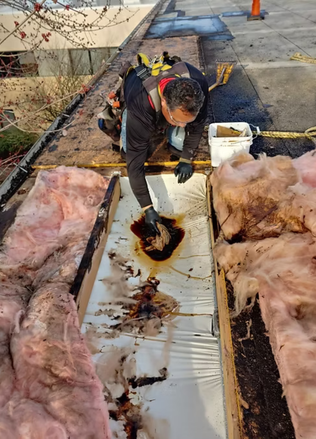 Worker on a rooftop repairing a damaged section beside pink insulation and a bucket of tools