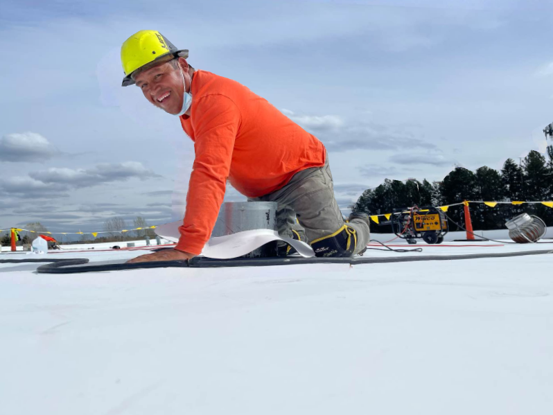 Worker in an orange shirt and hard hat kneeling on a white rooftop under construction.