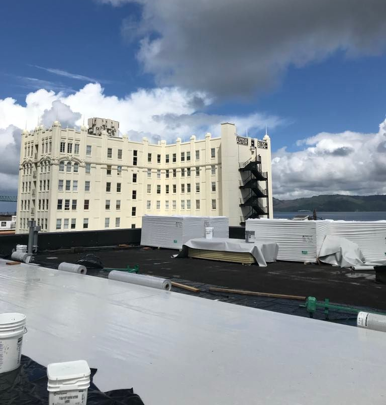 Rooftop with white roofing material and buckets overlooking a cream-colored building under a cloudy blue sky