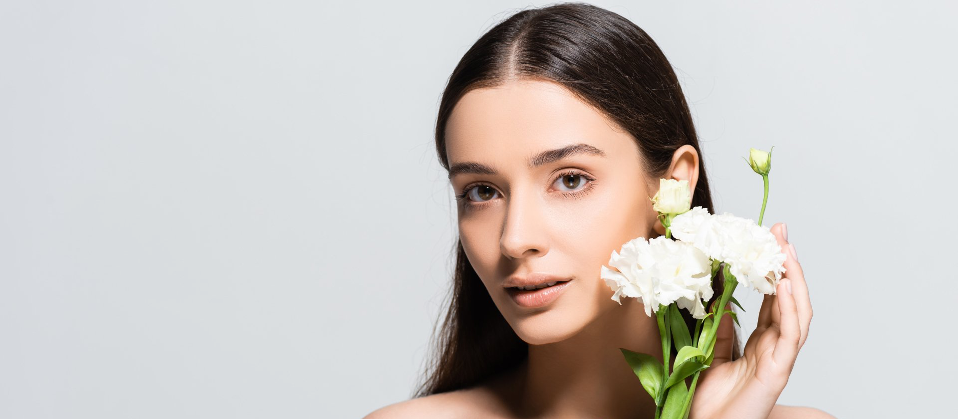 Eine Frau mit langen dunklen Haaren hält vor grauem Hintergrund weiße Blumen vor ihr Gesicht.