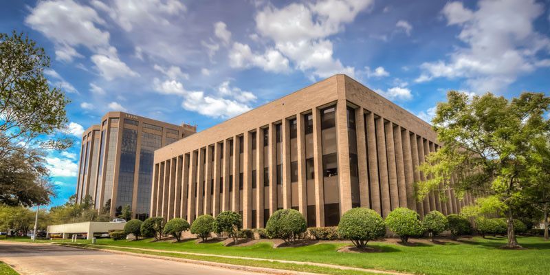 Two large beige buildings with many windows under a blue sky with fluffy clouds. Green grass and trees in front.