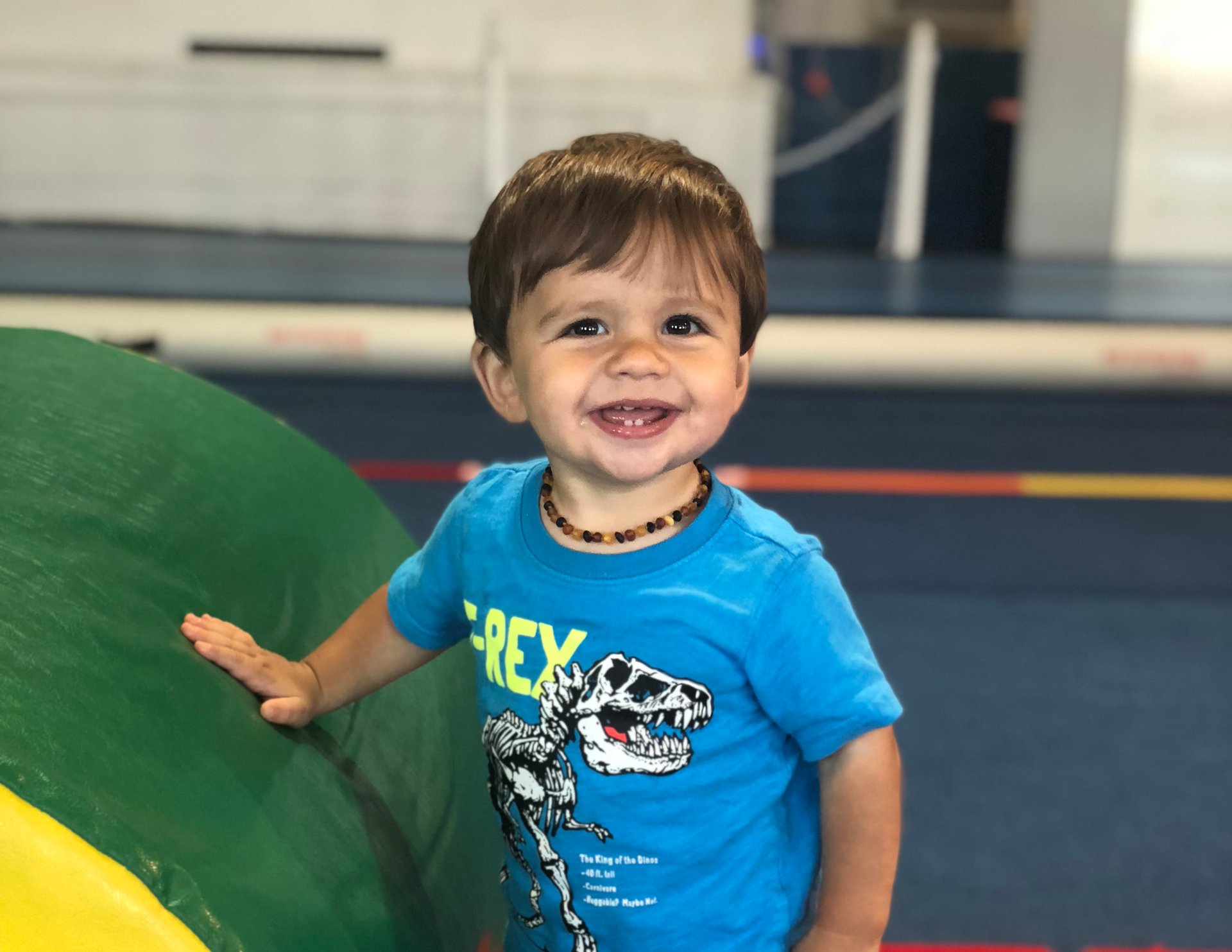 Smiling child wearing a blue t-shirt with a dinosaur design, leaning on a green ball in a gymnasium.