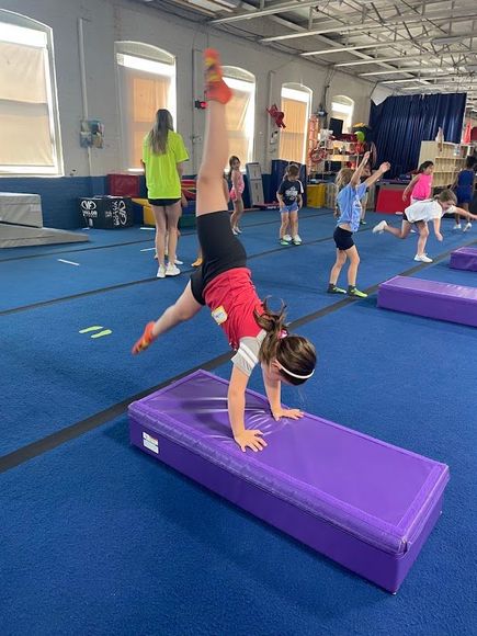 Girl doing a handstand on a purple mat in a gymnastics gym. Other children are visible in the background.