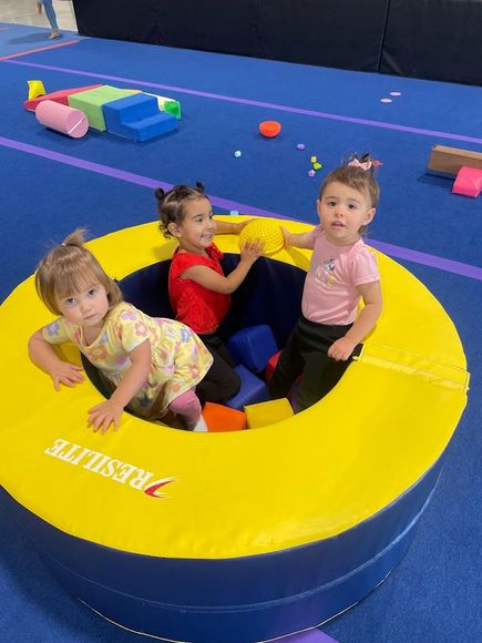 Three young children in a yellow ball pit on a blue gym floor. One child smiles, others reach for balls.