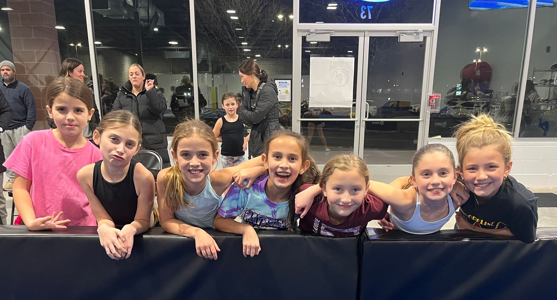 Group of young girls smile at the camera, leaning on a padded barrier in a gym.