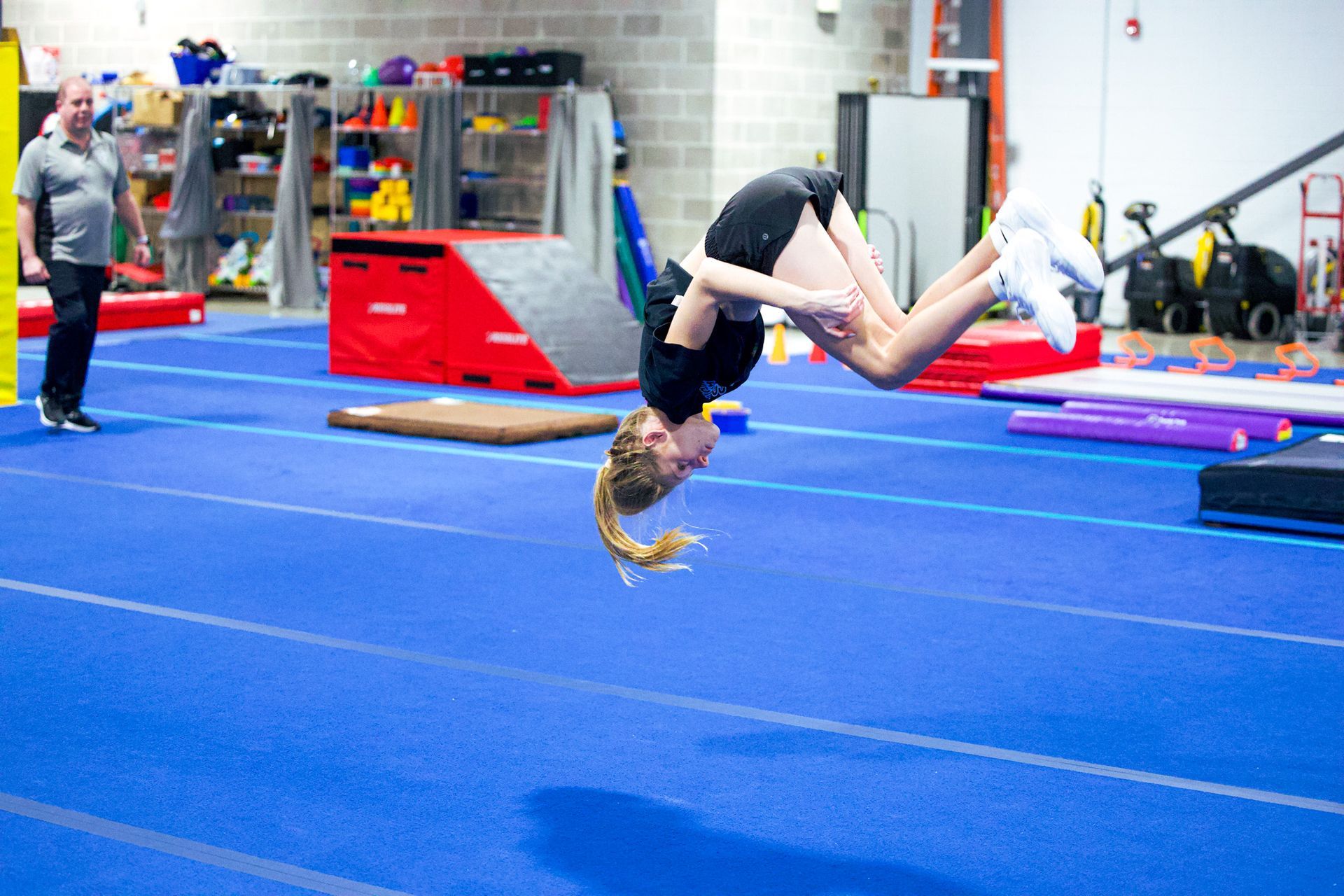 Gymnast in a purple leotard balances on a beam, arms raised, against a black background.