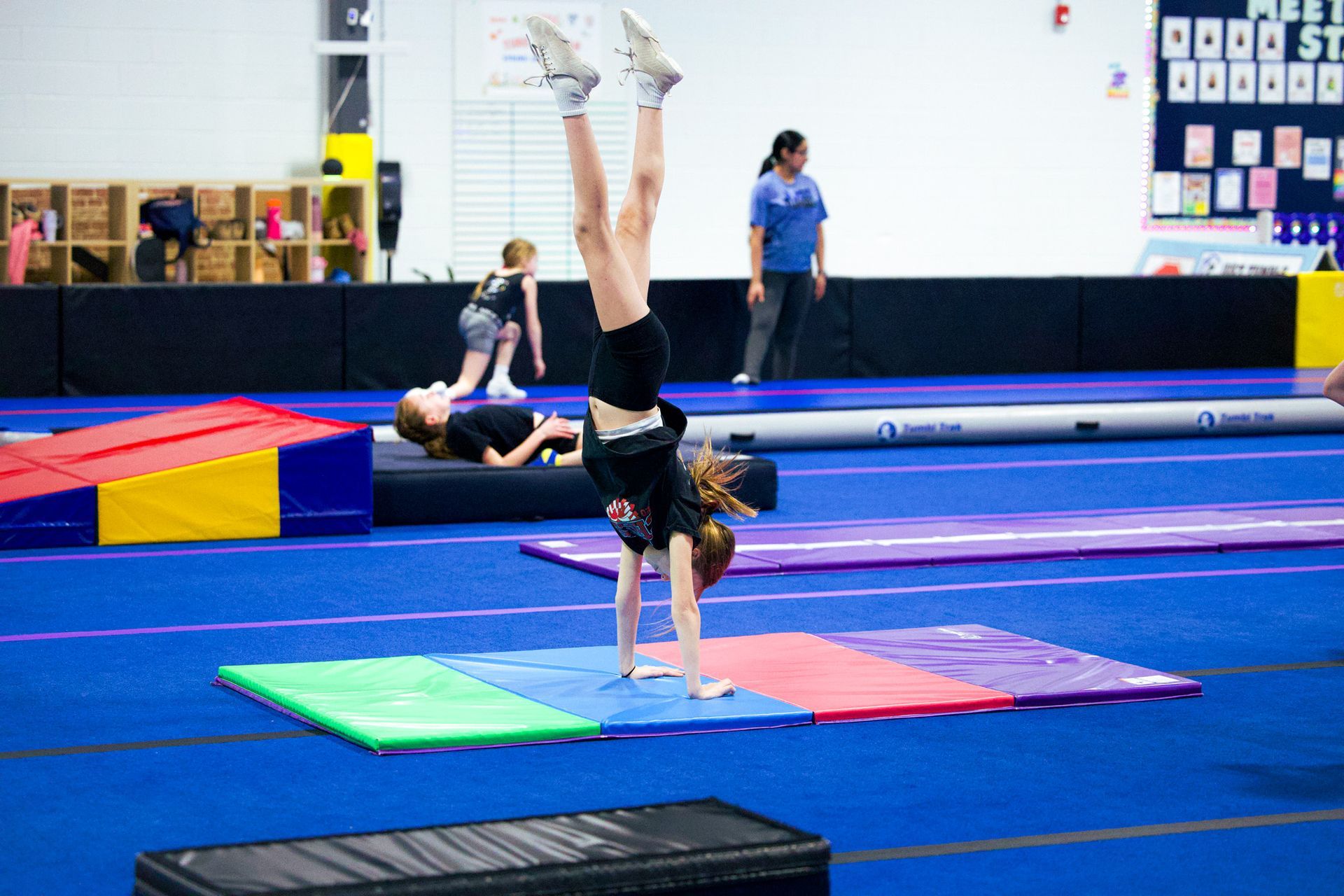 Girl doing a handstand on a blue mat, legs split in a gymnastics studio.