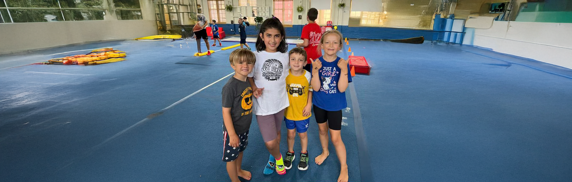Four children smiling, posing on a blue surface, inside a large building. Others are in the background.