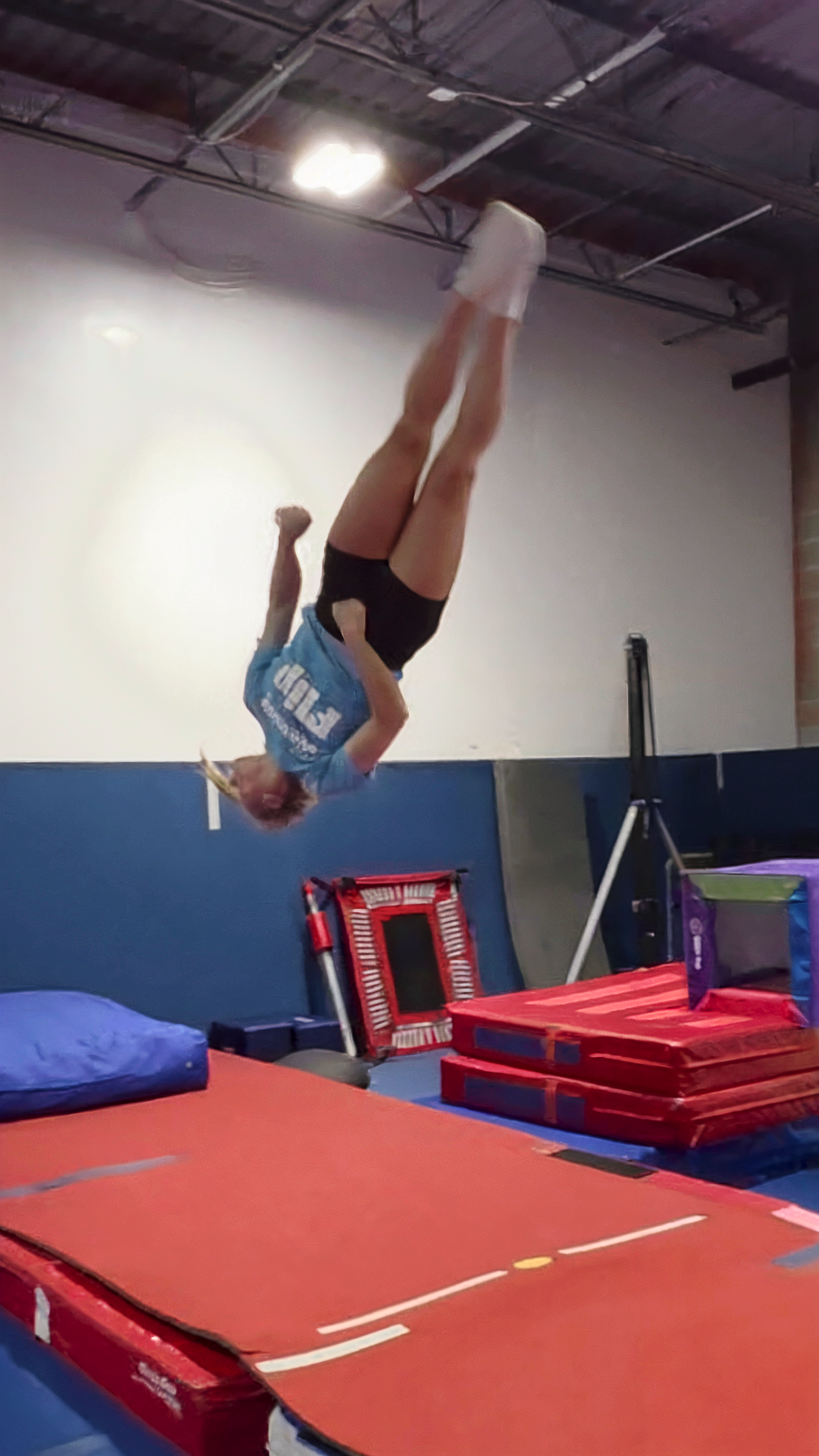 Gymnast in mid-air backflip, blue shirt, black shorts, white socks, red mat, gym setting.