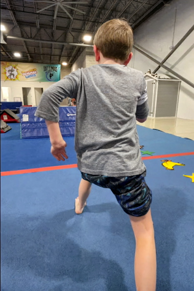 A child in grey shirt and patterned shorts stands on a blue mat, arm extended. Indoors.