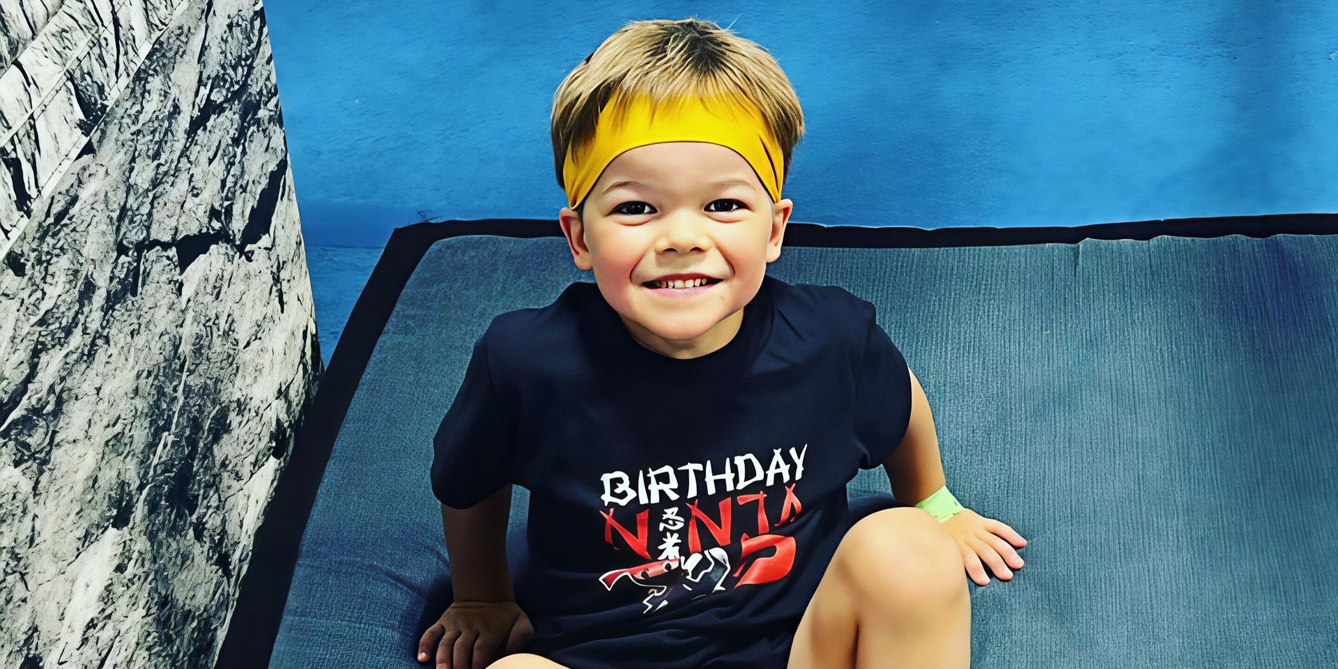 A smiling child wearing a crown and "Birthday Ninja" shirt sits on a padded surface. Blue background.