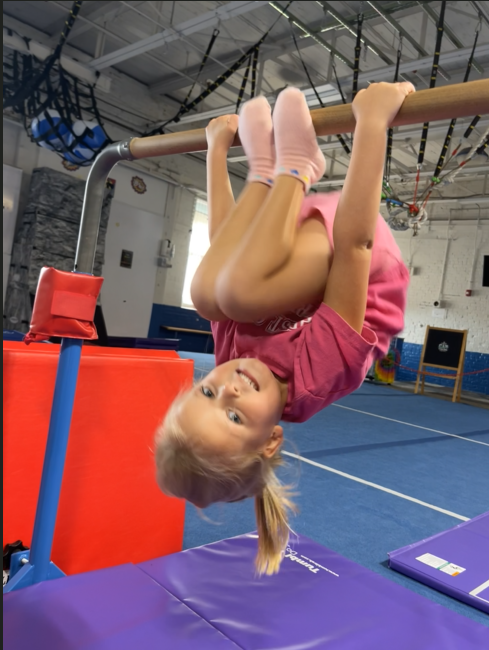 Girl hanging upside down on a horizontal bar in a gym, wearing pink and smiling.