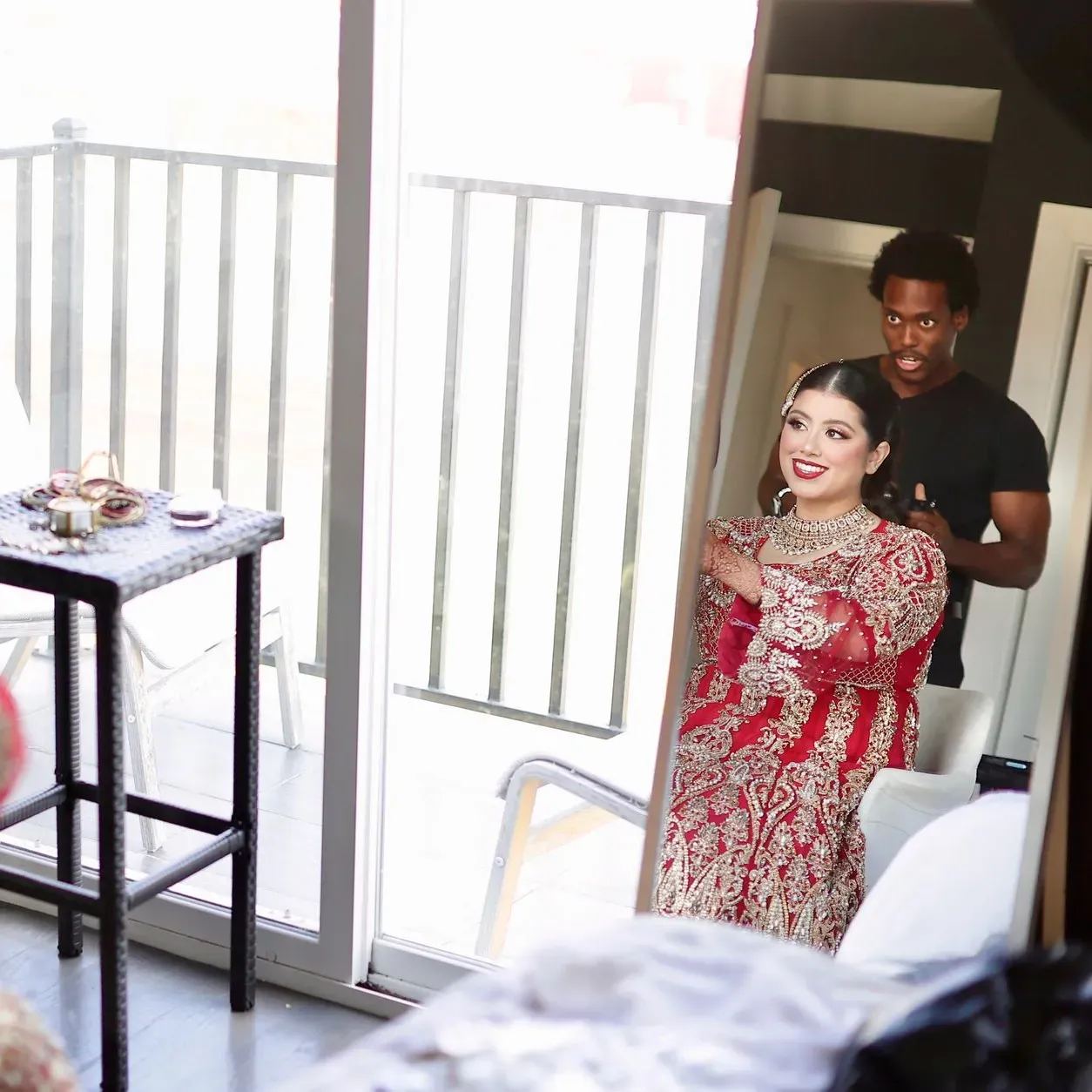 Woman in a red patterned dress posing in a bright hallway, with a man standing behind her near a doorway.