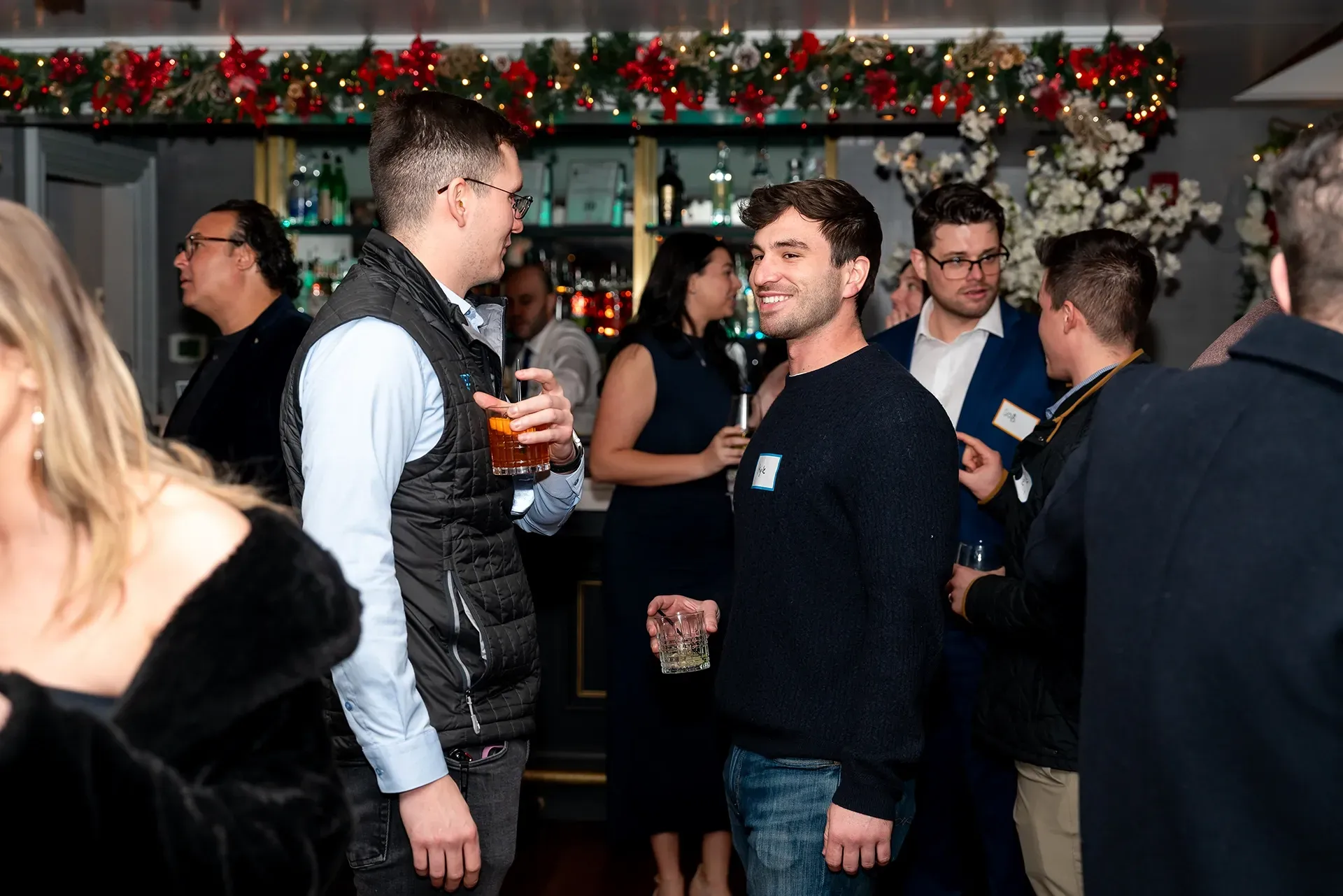 People mingling at a festive indoor holiday party, with garlands and red decorations overhead.