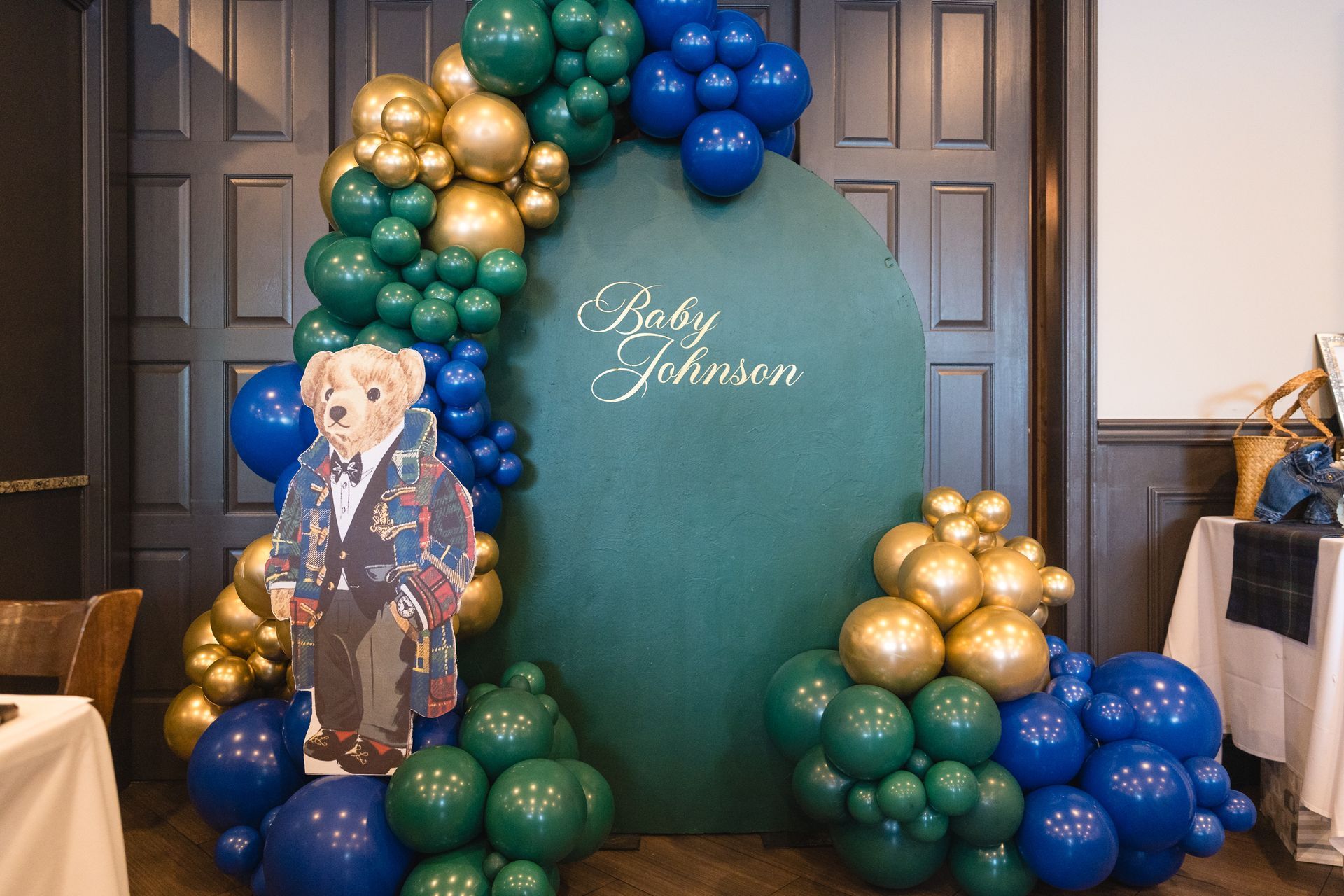 Balloon display in teal, gold, and blue around a teal sign with a teddy bear cutout at a party entrance