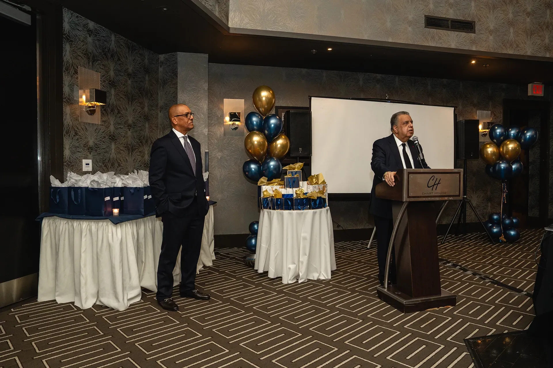Man speaking at a podium in a decorated banquet room, with another man standing nearby and tables with balloons.