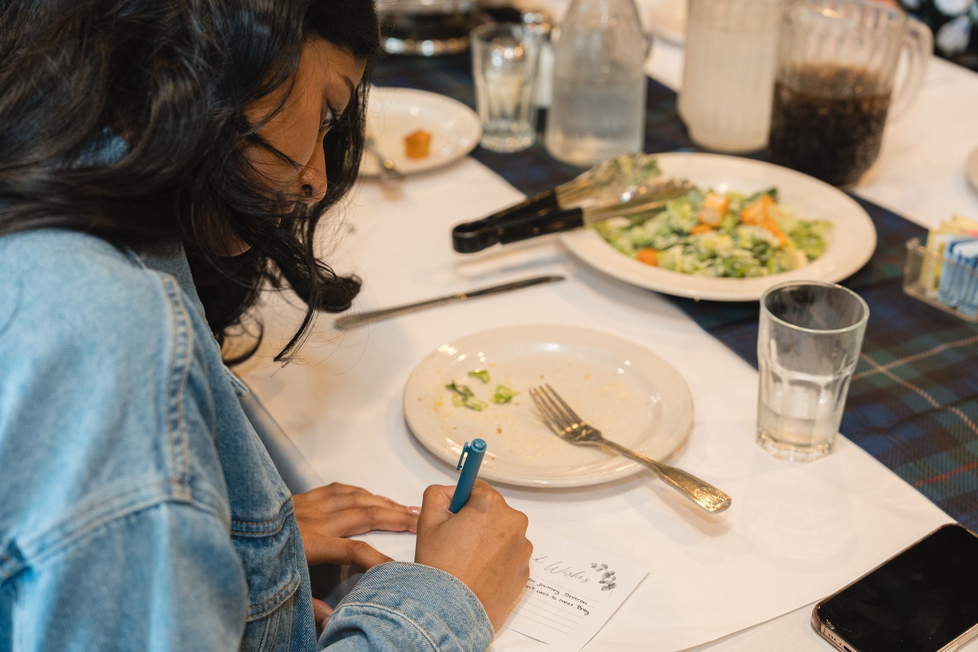 Person writing at a restaurant table with plates, drinks, and a meal in front of them