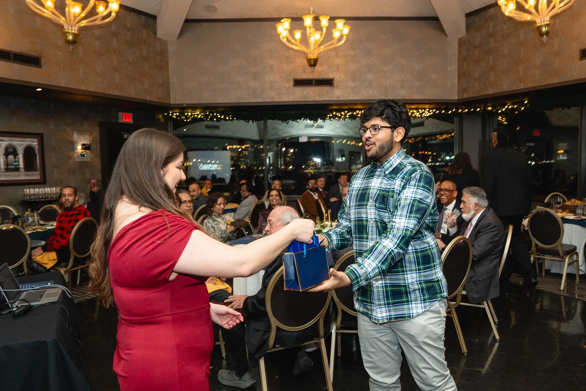 Two people exchanging an item at a formal indoor event with seated guests and chandeliers in the background
