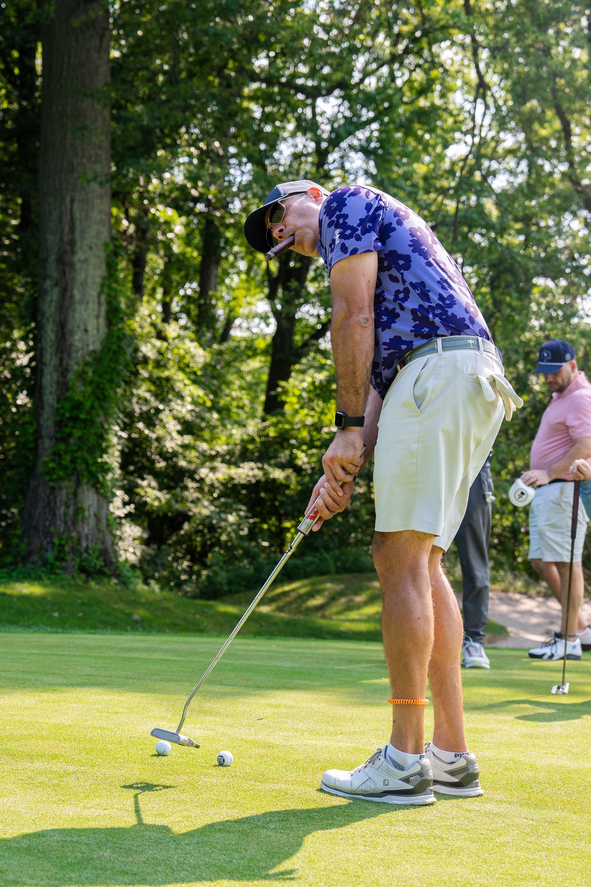 Golfer putting on a sunlit green, with trees in the background and another player behind.