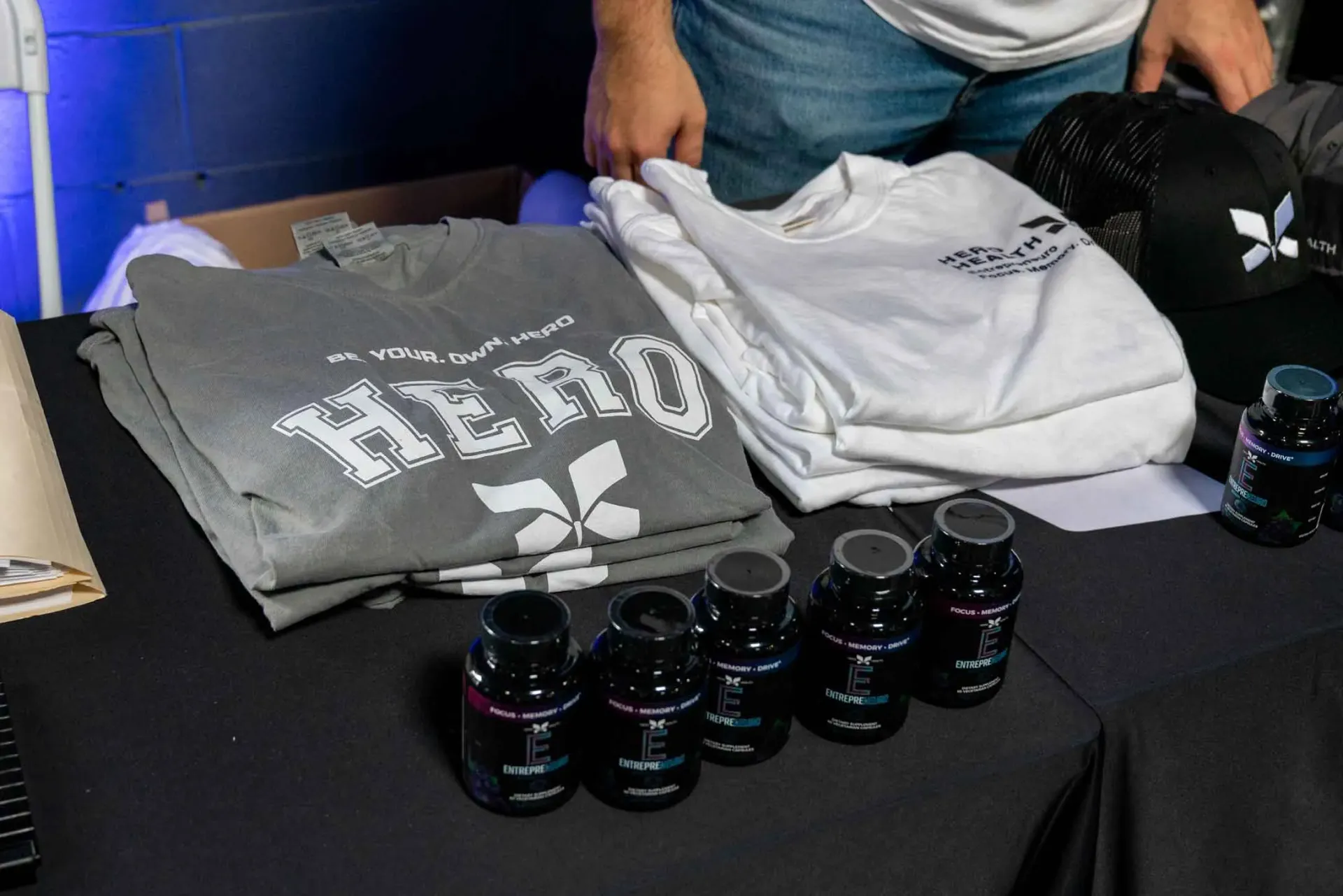A man is standing next to a table with shirts and bottles of pills on it.