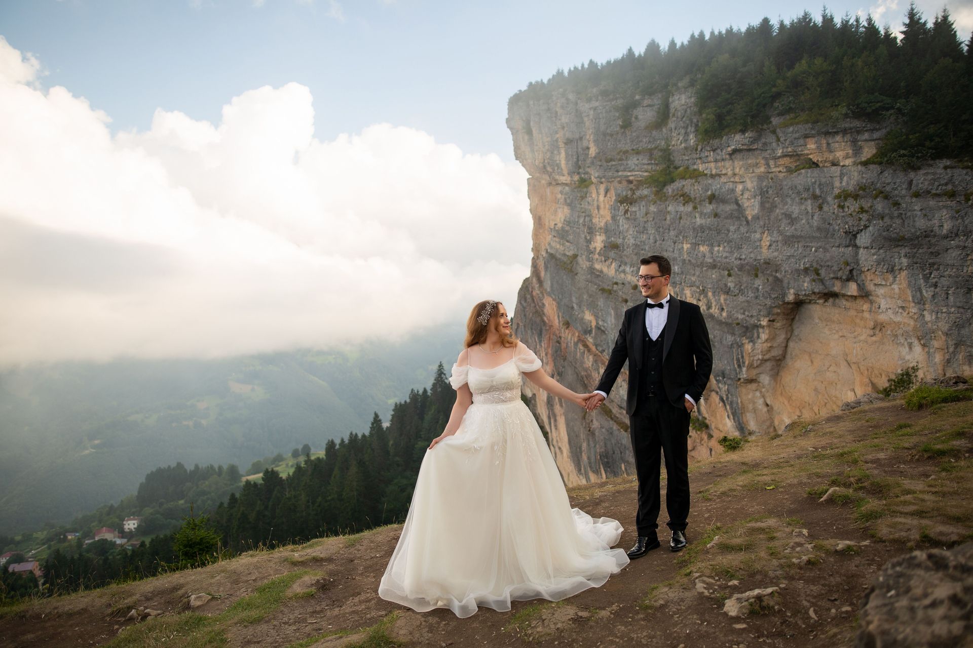 Bride and groom holding hands on a cliffside overlook, with mountains and clouds in the background