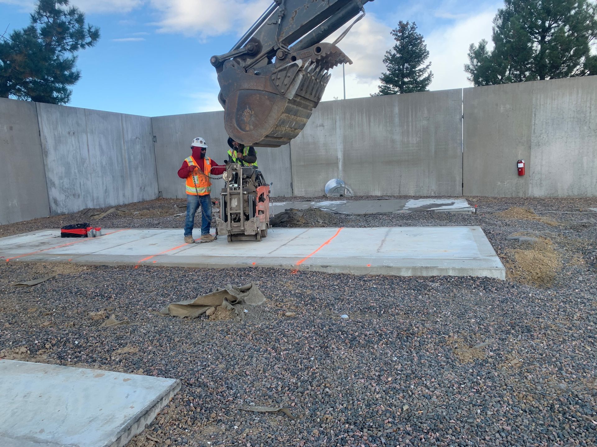 Person in orange overalls and gloves using a concrete saw on pavement.