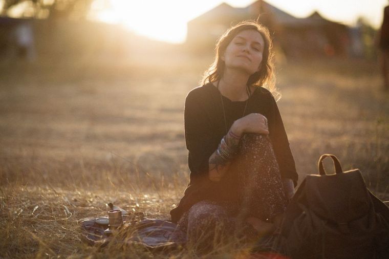 Woman sitting in a sunlit field at sunset, holding fabric beside a bag
