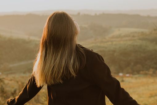 Person with long hair facing a hazy sunset over rolling hills, arms outstretched
