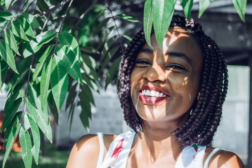 Smiling person in striped top under green leaves, sunlight casting dappled shadows across face