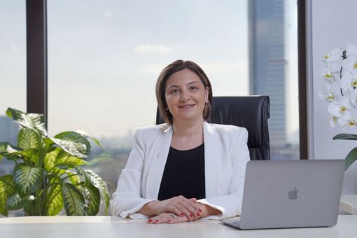 Woman seated at a desk in an office, smiling beside a laptop and green plant.